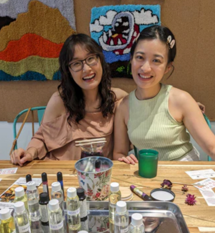 Two people sit closely together and smile. They are sitting at a table with candle making supplies on it. There are tufted wall hangings behind them