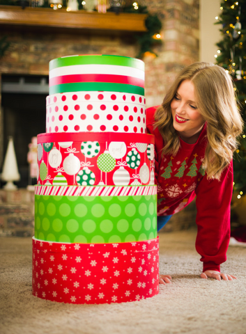A giant gift wrapped in green, red, and white sits on a white carpet. A person wearing a Christmas sweater peeks around from behind the tower of gifts. There is a Christmas tree and a fireplace in the background