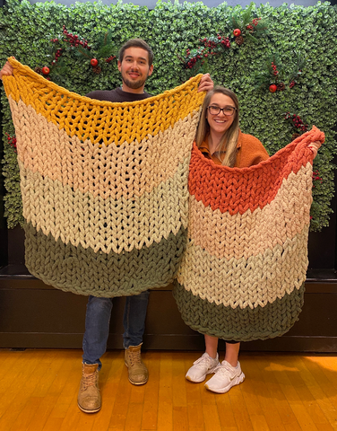 Two people smile and hold up hand made blankets in yellow, white, green, and orange. They stand in front of a moss wall