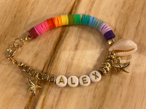 A hand-made beaded bracelet with rainbow beads, shells, and beads spelling out the name "Alex." The bracelet sits on a wood background
