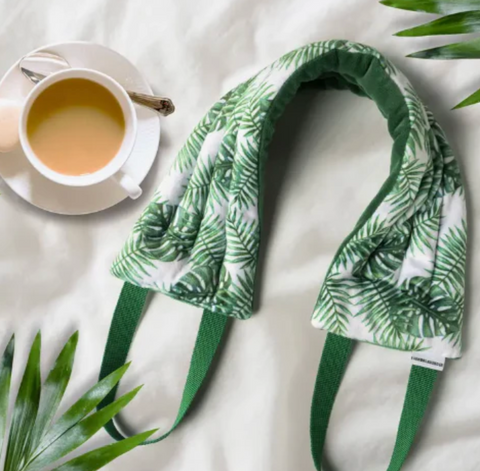 A leaf-patterned neck wrap laying on a white bed spread next to a cup of tea