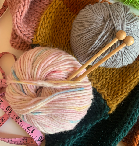 Two balls of yarn and a pair of knitting needles lying on top of a knitting fabric. A tape measure can be seen poking out from the fabric.