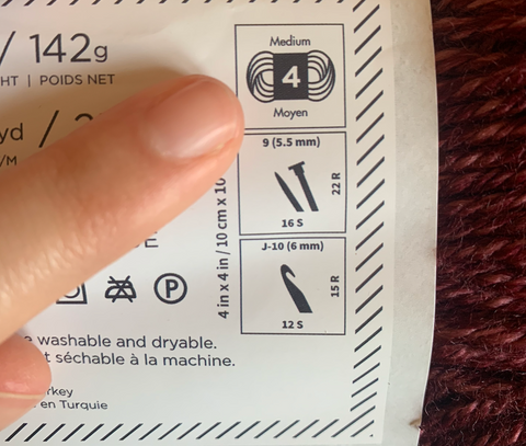 A finger pointing to the Medium Size 4 yarn weight label on the ball band of a ball of yarn
