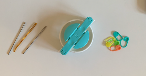 From left to right, three tapestry needles, a pom pom maker, and a pile of five stitch markers.