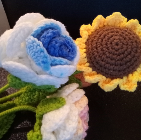 A crochet blue rose, a crochet sunflower, and a pink crochet rose on a black background.