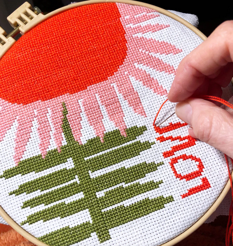 An embroidery hoop holding a work-in-progress cross-stitch piece. The image is a red and pink flower with a green stem, with "Love" written next to it. A hand is stitching the "Love".