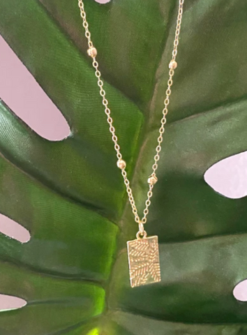 A gold necklace with an olive leaf pendant in front of a large green leaf.