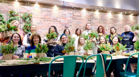 A group of people holding hand-crafted floral arrangements smiling. They stand behind a table full of crafting materials.