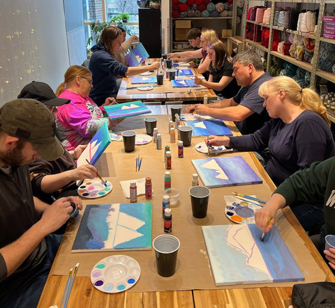 A group of people sit at a long table painting. Each person is working on a blue landscape painting with mountains. Behind them are shelves filled with crafting materials.