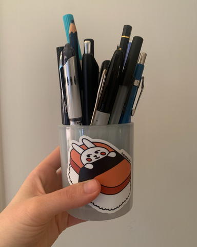 A jar decorated with a large vinyl sticker of a bunny in a sushi bed. There are pens and pencils in the jar.