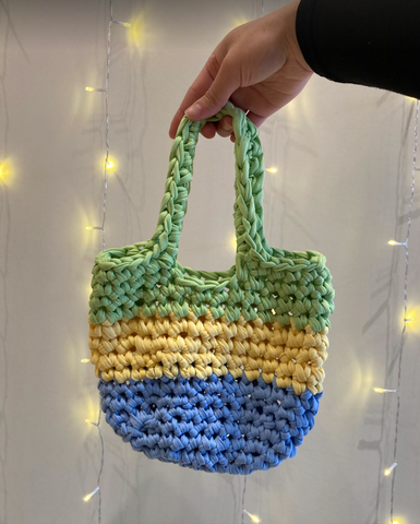 A green, yellow, and blue tote bag made out of t-shirt yarn and held against a white background.