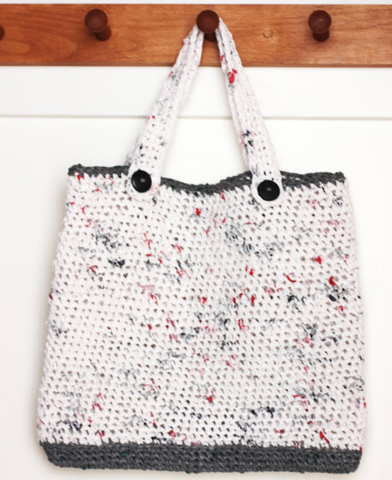 A white and black tote bag made out of plarn hanging from a wooden peg.