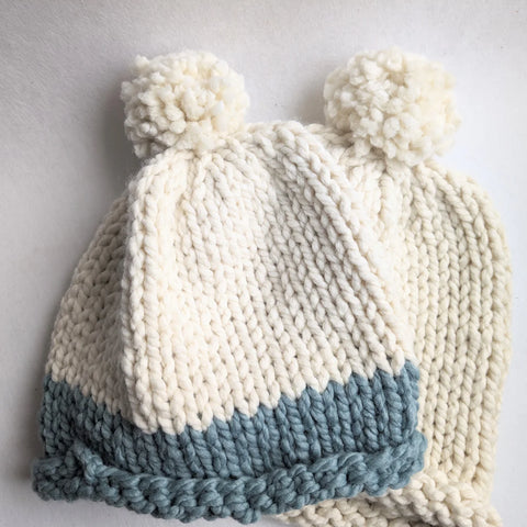 Two knit hats with pom poms laying one on top of the other. One is white with a mint green stripe and the other is all white.