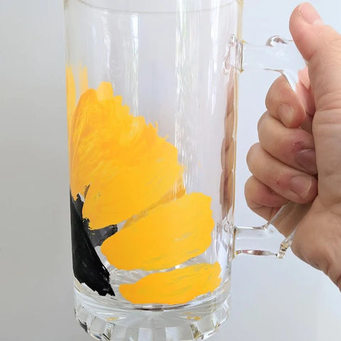 A glass mug with a sunflower painted on it held in front of a white background