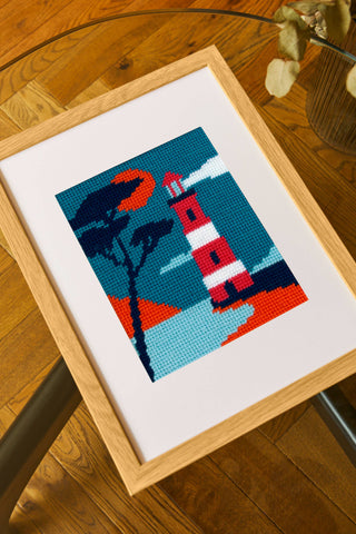 A needlepoint tapestry of a lighthouse, framed and lying on a wooden backdrop