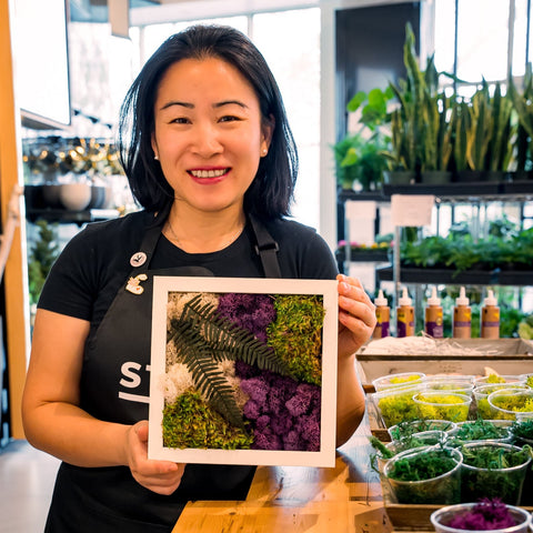 A woman holds up a piece of moss art in front of some plants