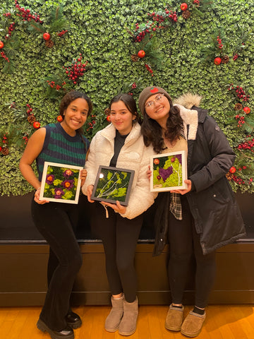 A group of three people standing in front of a moss wall holding moss art in their hands.