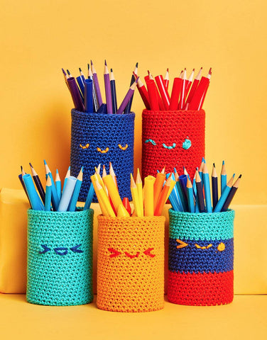 Five brightly colored pencil pots with pencils on them in front of a bright yellow background. Each pencil pot has an embroidered smiley face on it.
