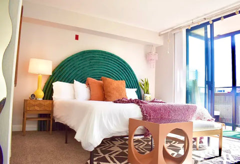 A shot of a bedroom with a pool noodle headboard covered in green velvet behind a bed with white bedsheets and brown-orange pillows.