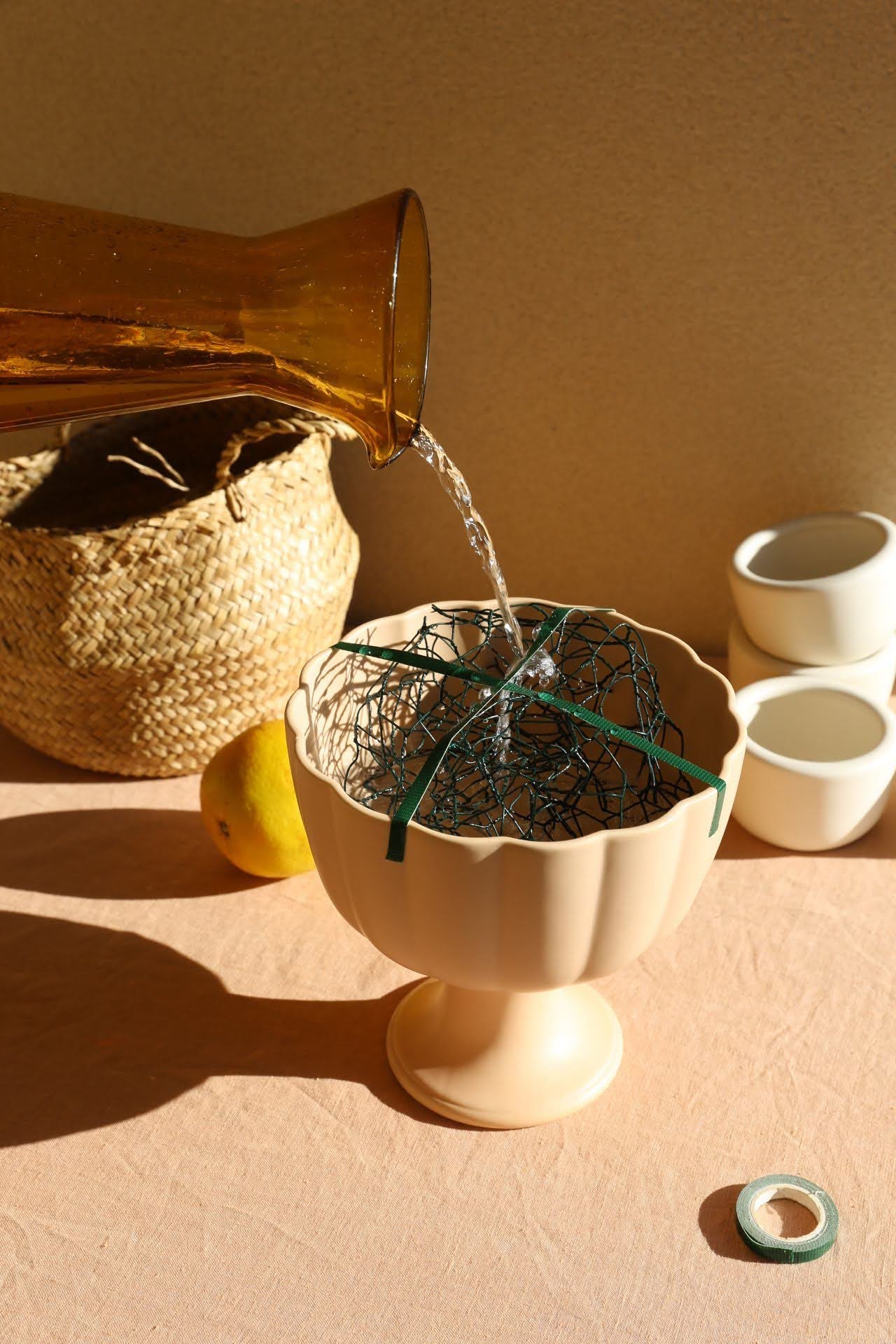 Light brown scallop edge footed bowl with chicken wire inside being filled with water from an amber glass jug