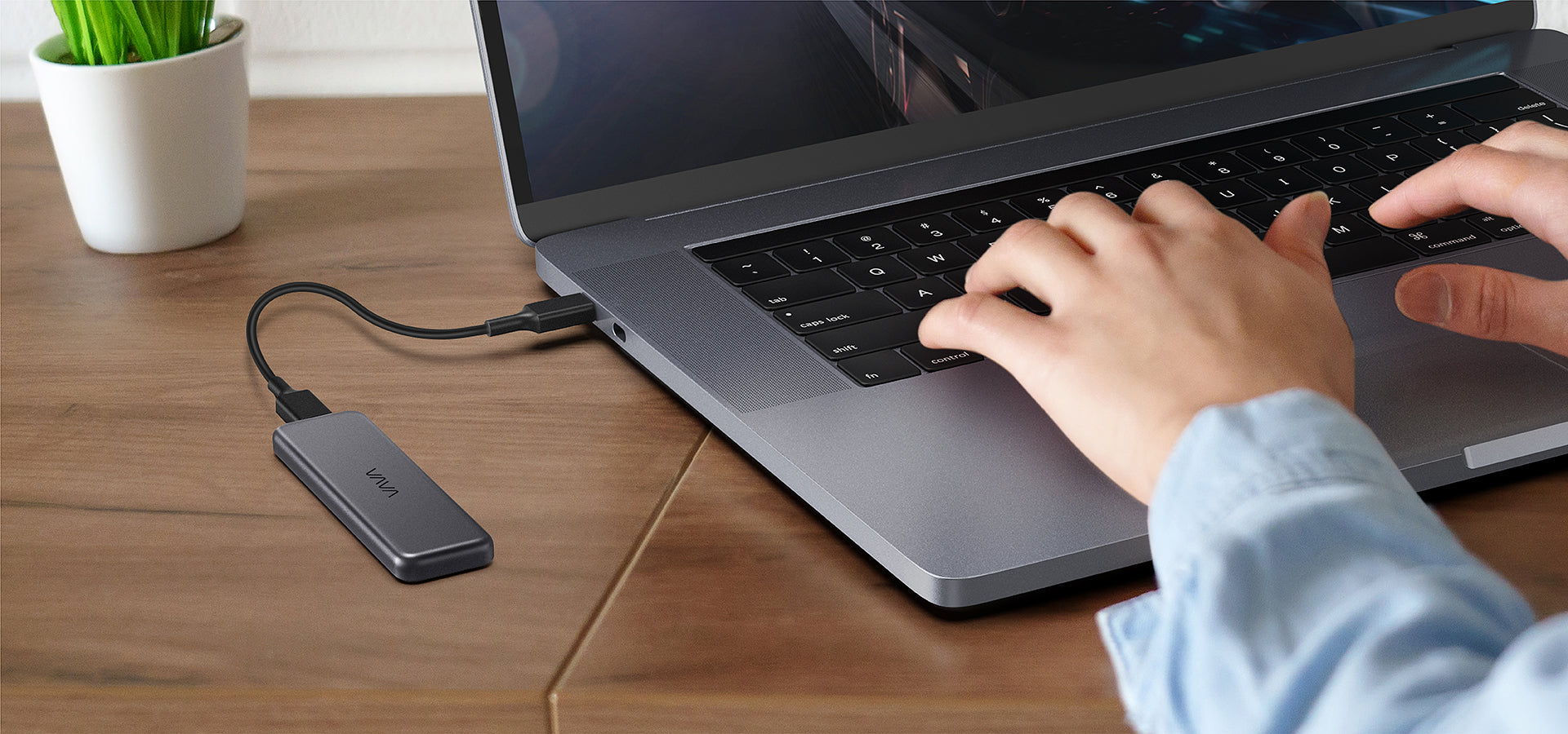 Hands typing on a laptop hooked up to a VAVA portable SSD on a brown wood desk