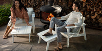 A couple relaxing around a fire pit in their white Neighbor adirondack chairs