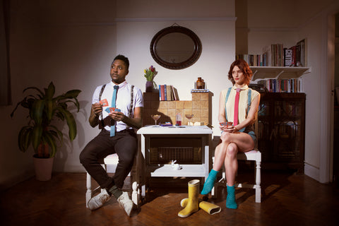 A man and woman sit holding playing cards next to a table, they are both wearing knitted suspenders and ties