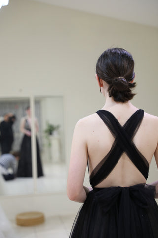 black cross back formal dress on brunette woman standing in front of mirror