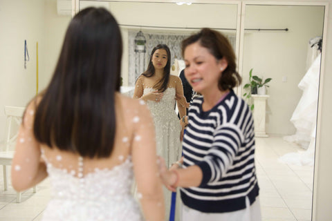 bridal designer fitting bride into her lace wedding dress in front of full length mirror