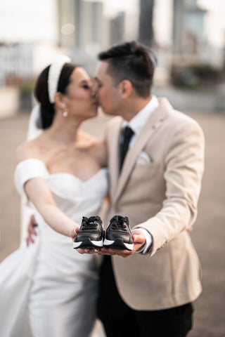 bride and groom kissing on their wedding day wearing wedding attire holding new born babys shoes
