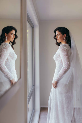 bride in her long sleeved lace wedding dress looking at herself in the mirror