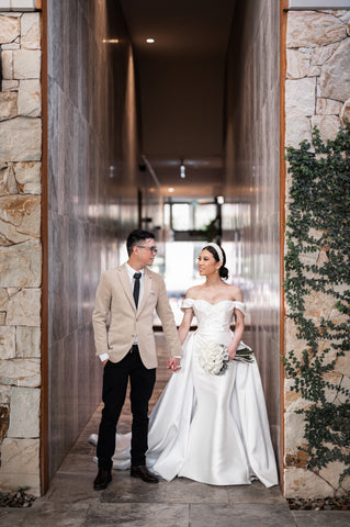 bride wearing white custom wedding dress holding hands with husband in suit in stone hallway