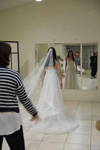 dressmaker in striped blue and white shirt laying out the veil worn by a bride in her wedding dress standing in front of studio mirrors while a woman in black photographs them