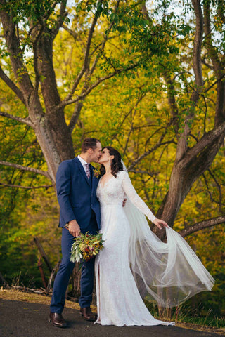 groom in blue suit holding brides bouquet while she holds her wedding veil during their first kiss as a married couple in front of green and yellow trees in a forrest