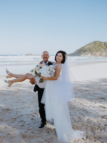 husband carrying wife on beach in wedding attire