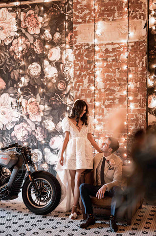 husband on seat and wife standing next to a motorcycle against a wall