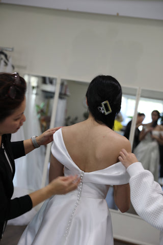 two women buttoning bride into her wedding dress in front of studio mirrors