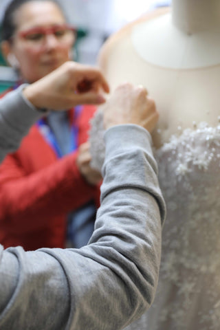 two women hand sewing lace cut outs onto a wedding dress displayed on a mannequin