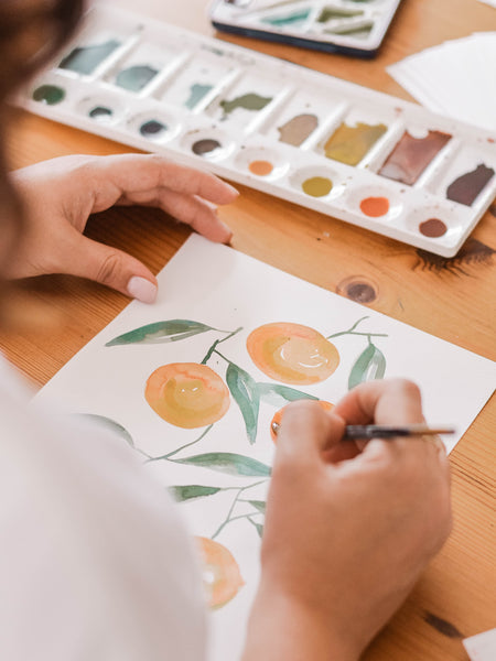 A lady painting an orange fruit with green leaves on a white paper