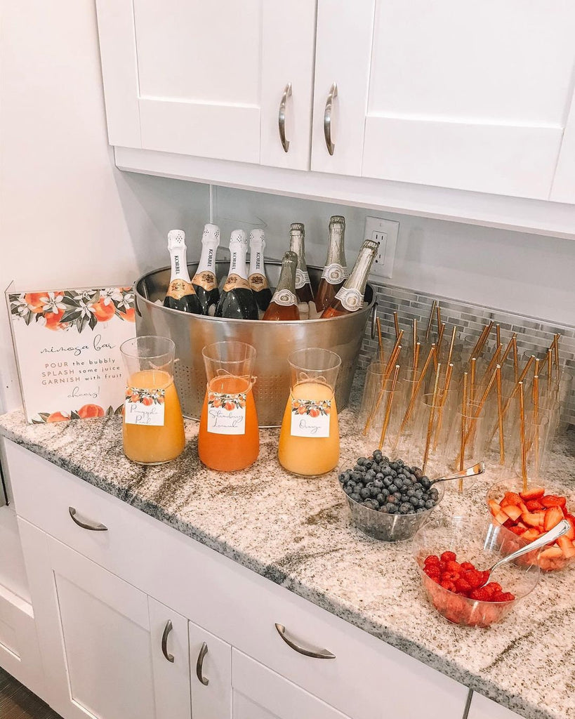 the perfect mimosa bar set up with watercolor peach signage and tags. cute glass champagne flutes with golden straws and a champagne bucket with several bowls of fruit on the side.