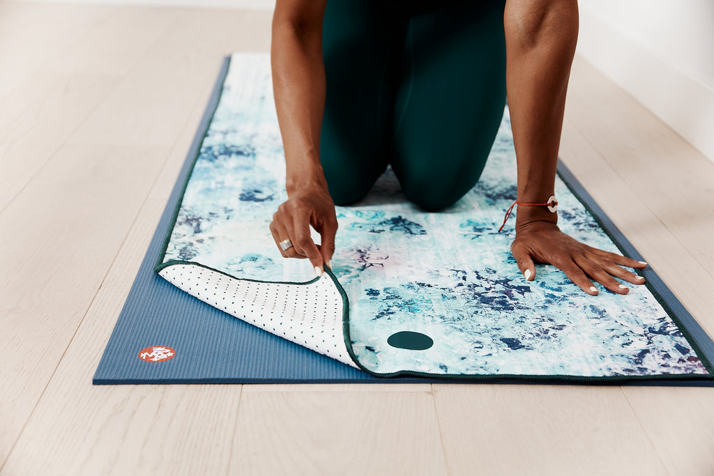 Yogi kneeling on her Yogitoes Mat Towel placed on her yoga mat lifting the corner of the towel to show the silicon nubs that grip the mat - Manduka | Eco Yoga Store