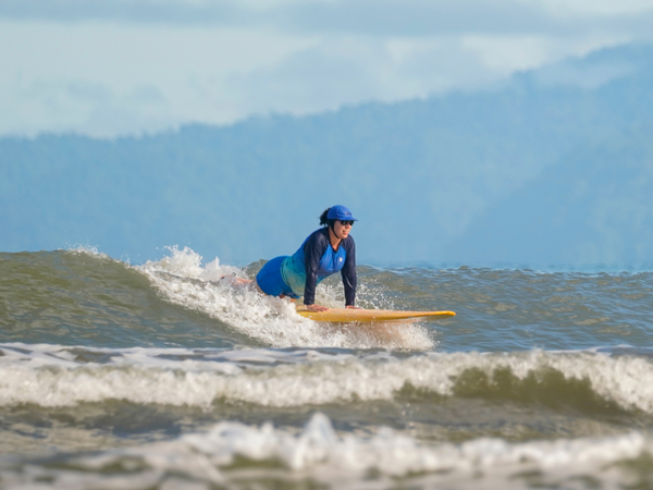 A surfer in a blue rashguard and hat rides a breaking wave, in a plank position on the board..