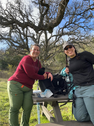 The image depicts two smiling female hikers sitting at a picnic table under a large oak tree. They appear to be taking a break during a hike, with one woman wearing a red long-sleeve shirt and green pants, and the other wearing a black long-sleeve shirt and gray pants. Hiking gear, including a backpack and water bottles, can be seen on the table. The surrounding landscape suggests they are enjoying a break in a natural setting during their outdoor adventure.