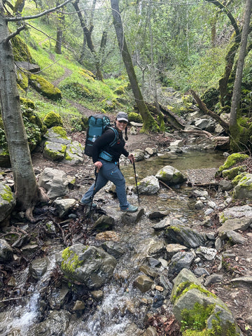 A hiker with a backpack and hiking poles carefully crosses a rocky stream bed in a lush forest. The trail follows the stream, which is lined with mossy boulders and surrounded by dense trees and foliage.