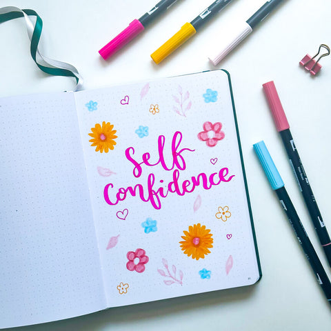 Un journal ouvert avec « confiance en soi » écrit en rose, entouré de gribouillages de fleurs