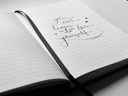 Quote 'You can learn to love yourself' written on a lined journal, in black and white