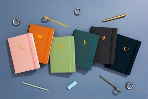 A collection of different coloured journals on a blue background, surrounded by stationery