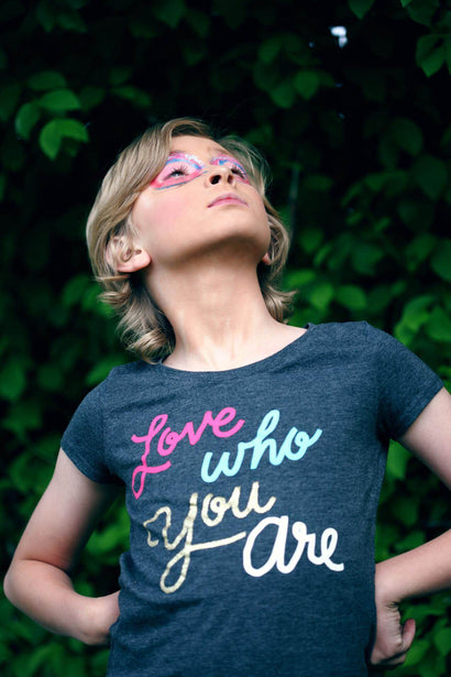 A child standing in a superhero pose in a t-shirt that reads 'love who you are'