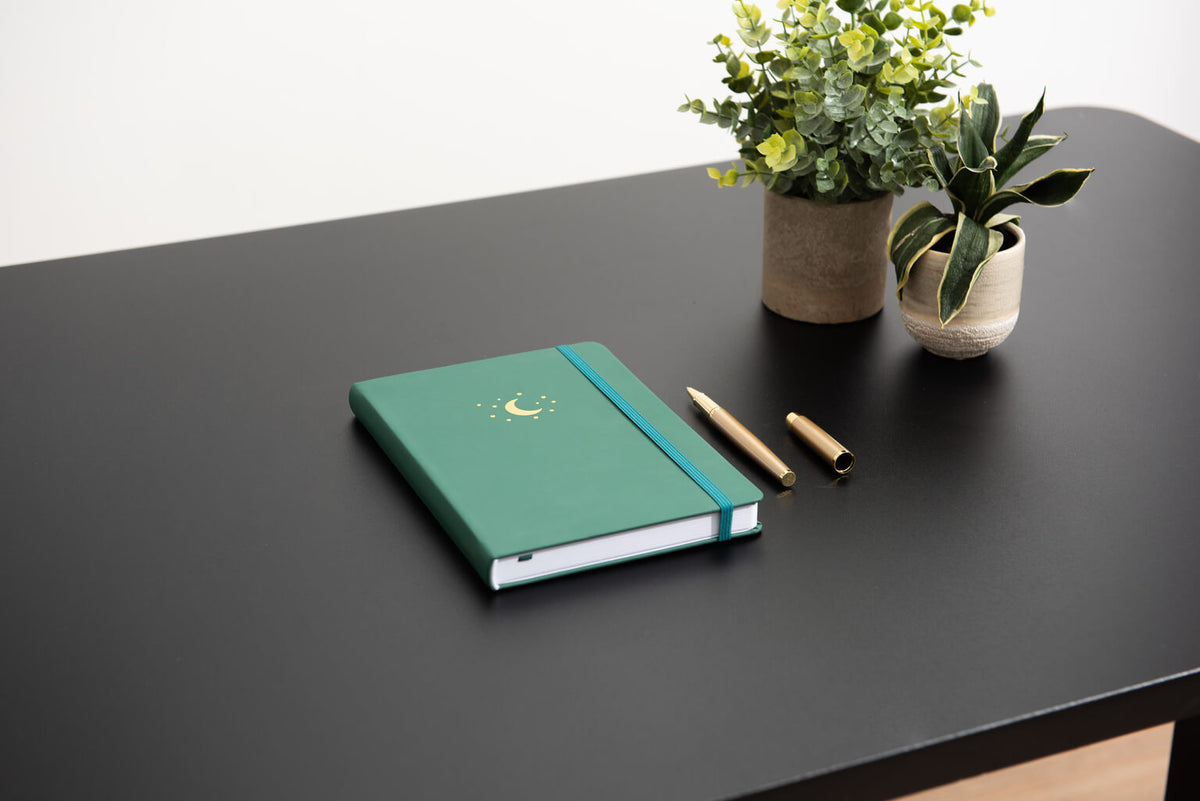 Forest green journal lying on a black desk beside plants and a gold pen