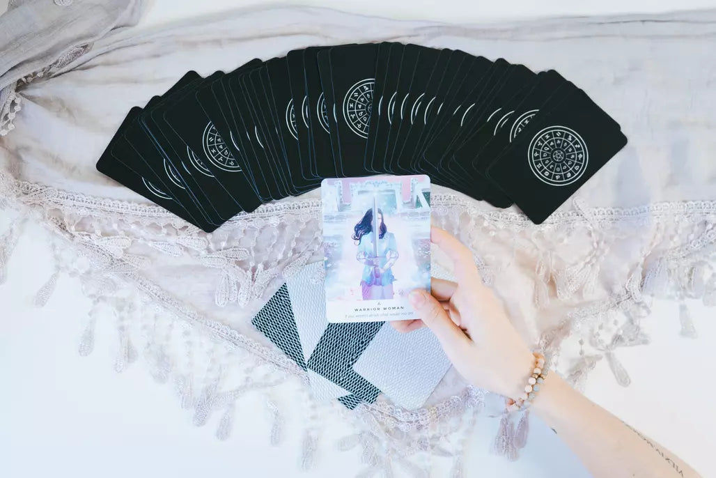 A deck of black tarot cards spread across a bed, with a hand holding one card